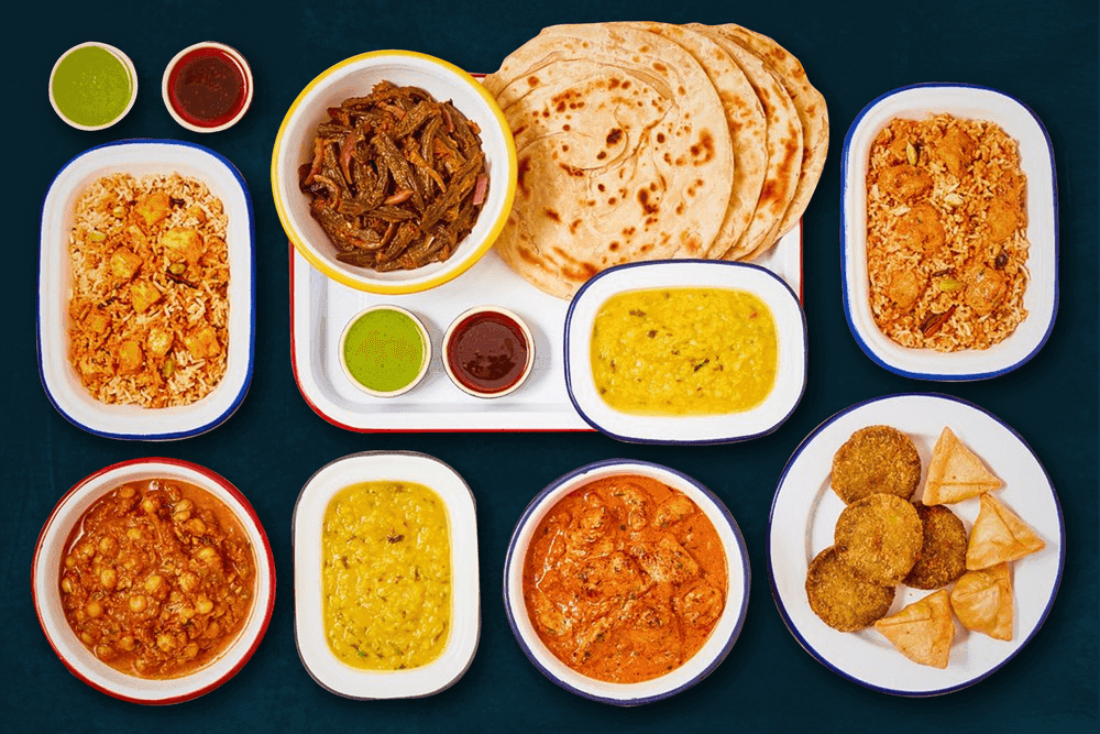 An assortment of authentic Indian dishes from General Bilimoria's Canteen, featuring butter chicken, dal, chana masala, biryani, crispy samosas, pakoras, parathas, and flavorful chutneys, served on traditional enamel plates.